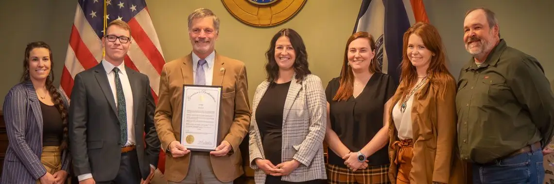 seven smiling people wearing formal attire stand in front of a U.S. flag and Wyoming state flag. The Wyoming governor holds a framed proclamation document with golden seal.