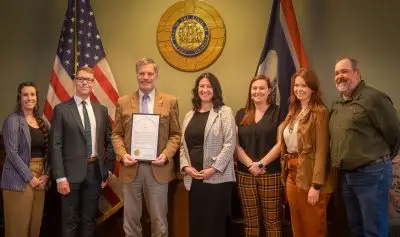 seven smiling people wearing formal attire stand in front of a U.S. flag and Wyoming state flag. The Wyoming governor holds a framed proclamation document with golden seal.