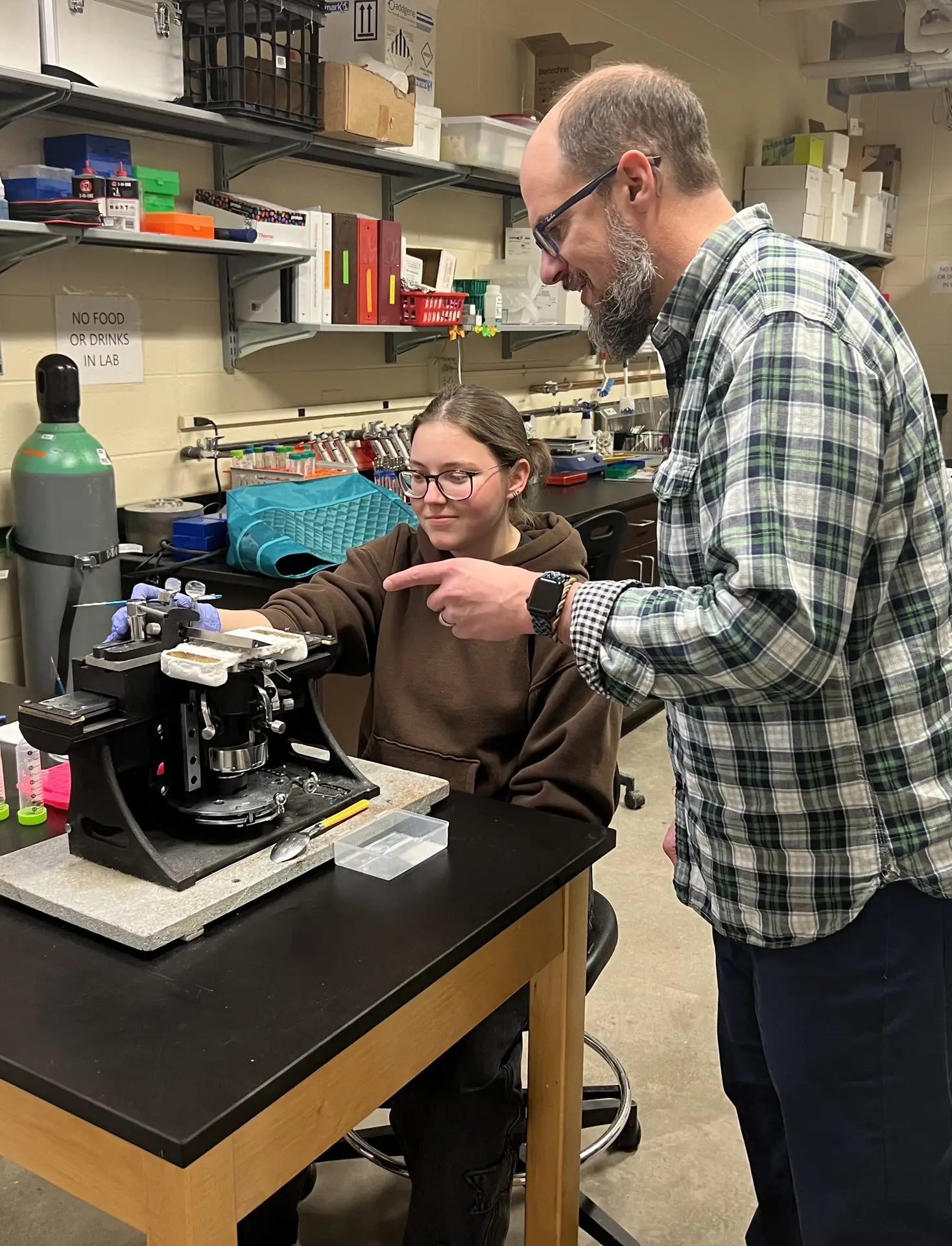 Man points at a scientific device on a table in front of a seated woman in a lab