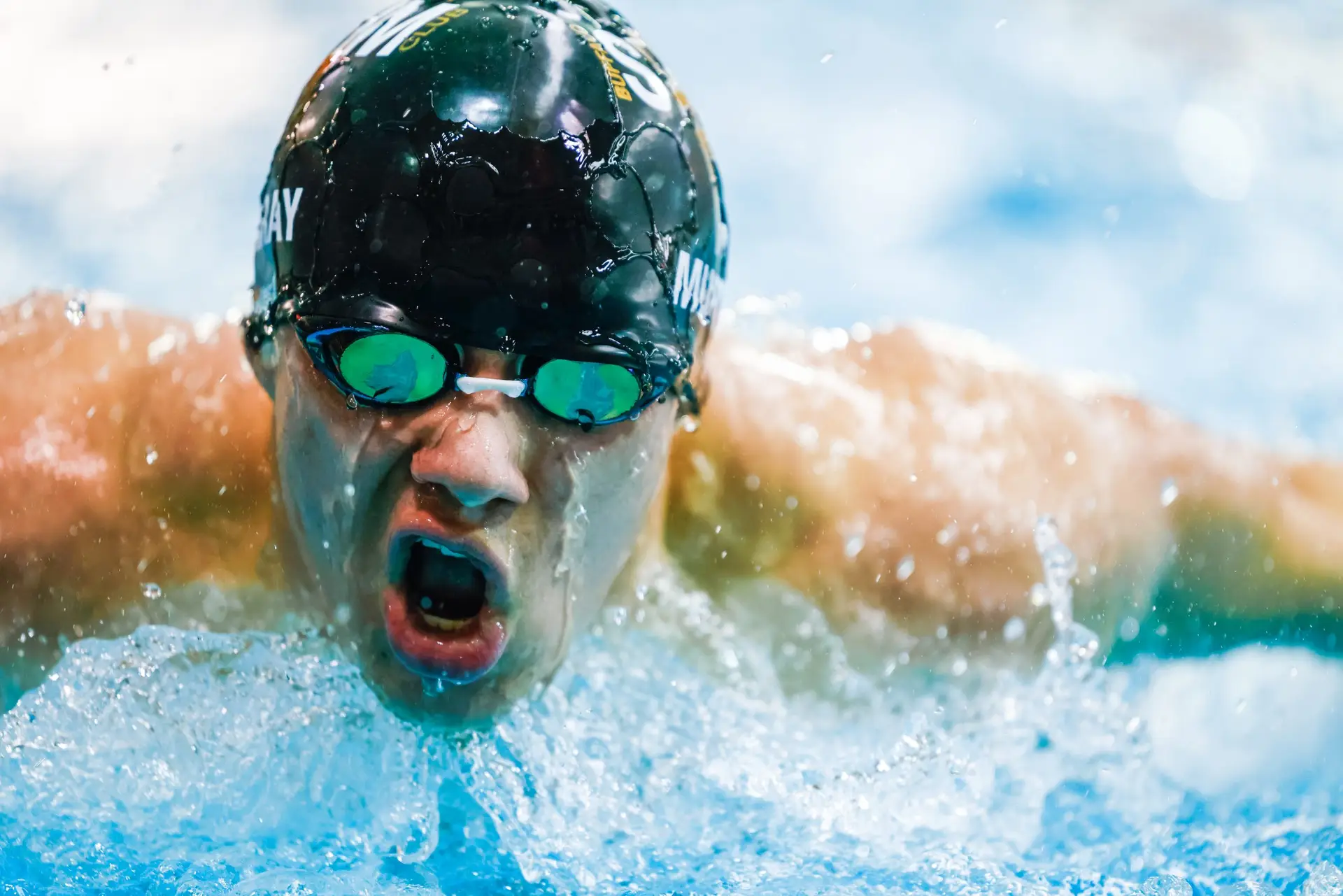 swimmer wearing black swim cap and green-tinted goggles moves through the water toward the camera at a high school swim meet