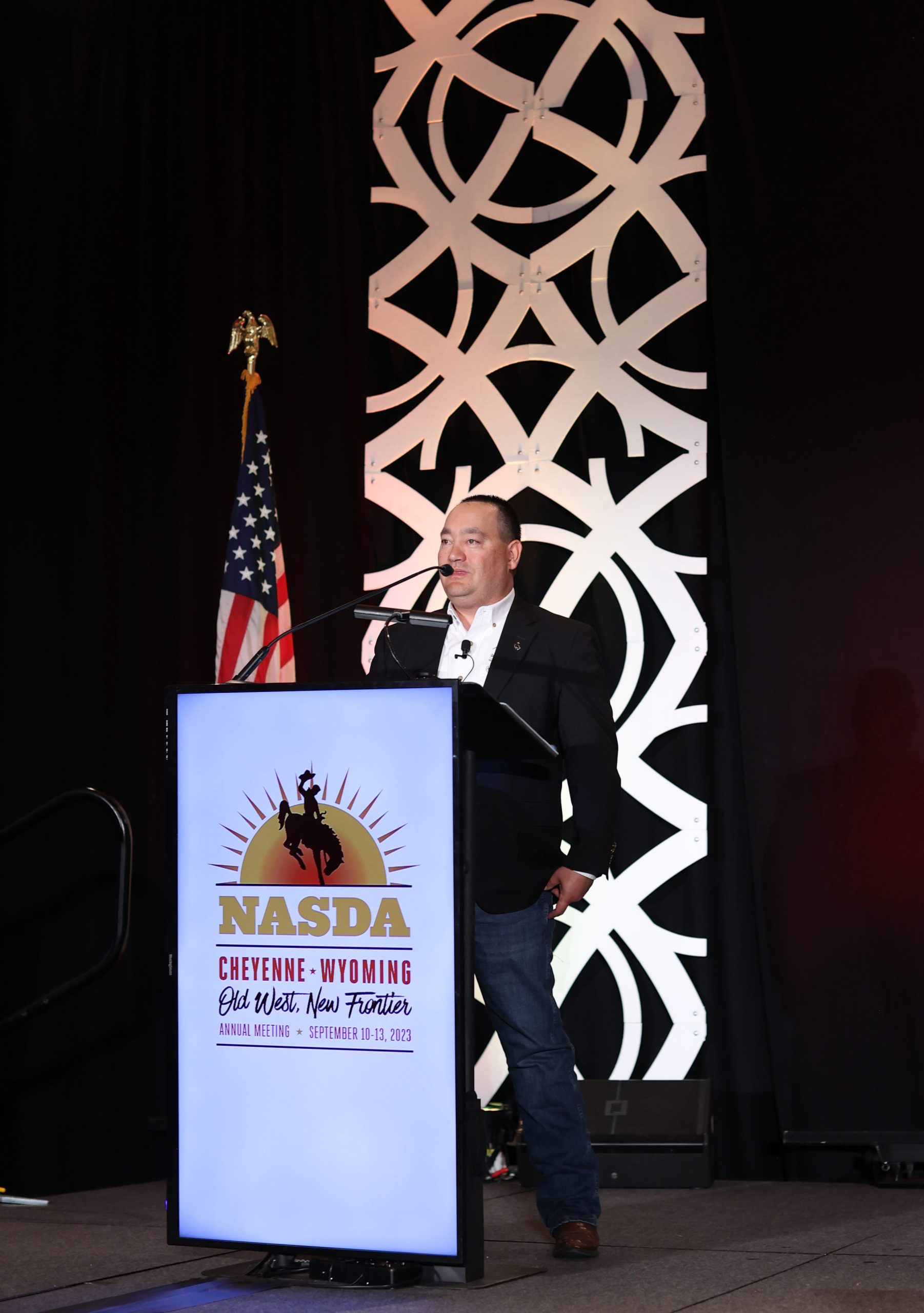 Man wearing formal attire stands behind a podium equipped with a mike and flanked by an American flag. A sign on the podium reads “NASDA Cheyenne Wyoming Old West, New Frontier Annual Meeting September 10-13, 2023”