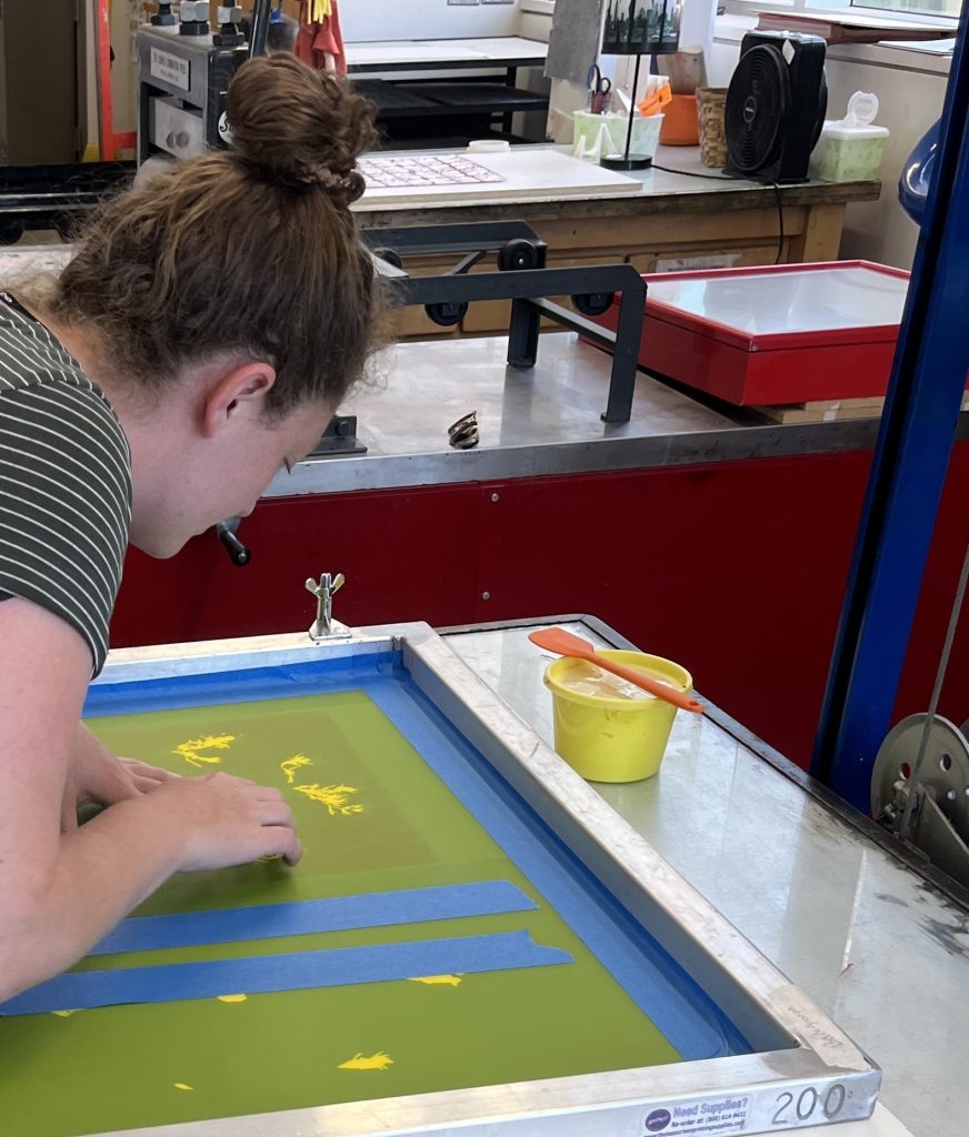 woman bends over a table, arranging a layer of a screen print with a green background and yellow drawings