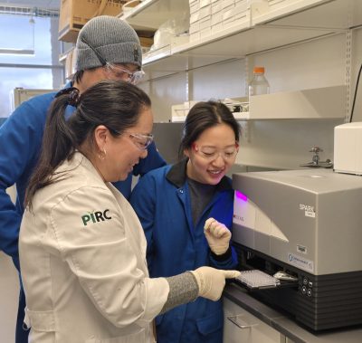 Three scientists wearing lab coats, safety glasses, and gloves gather around a scientific device being loaded with a small plastic tray