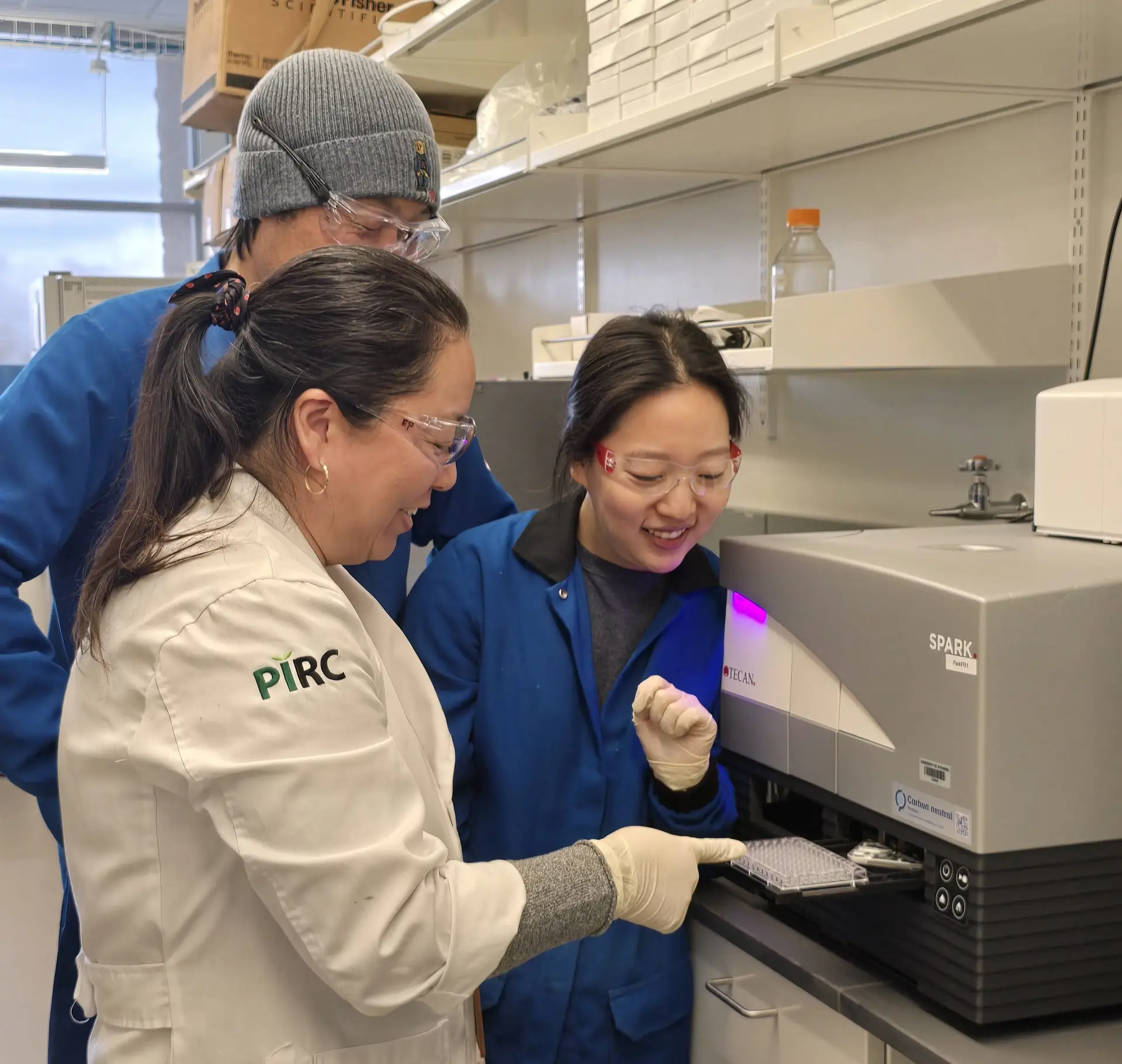 Three scientists wearing lab coats, safety glasses, and gloves gather around a scientific device being loaded with a small plastic tray