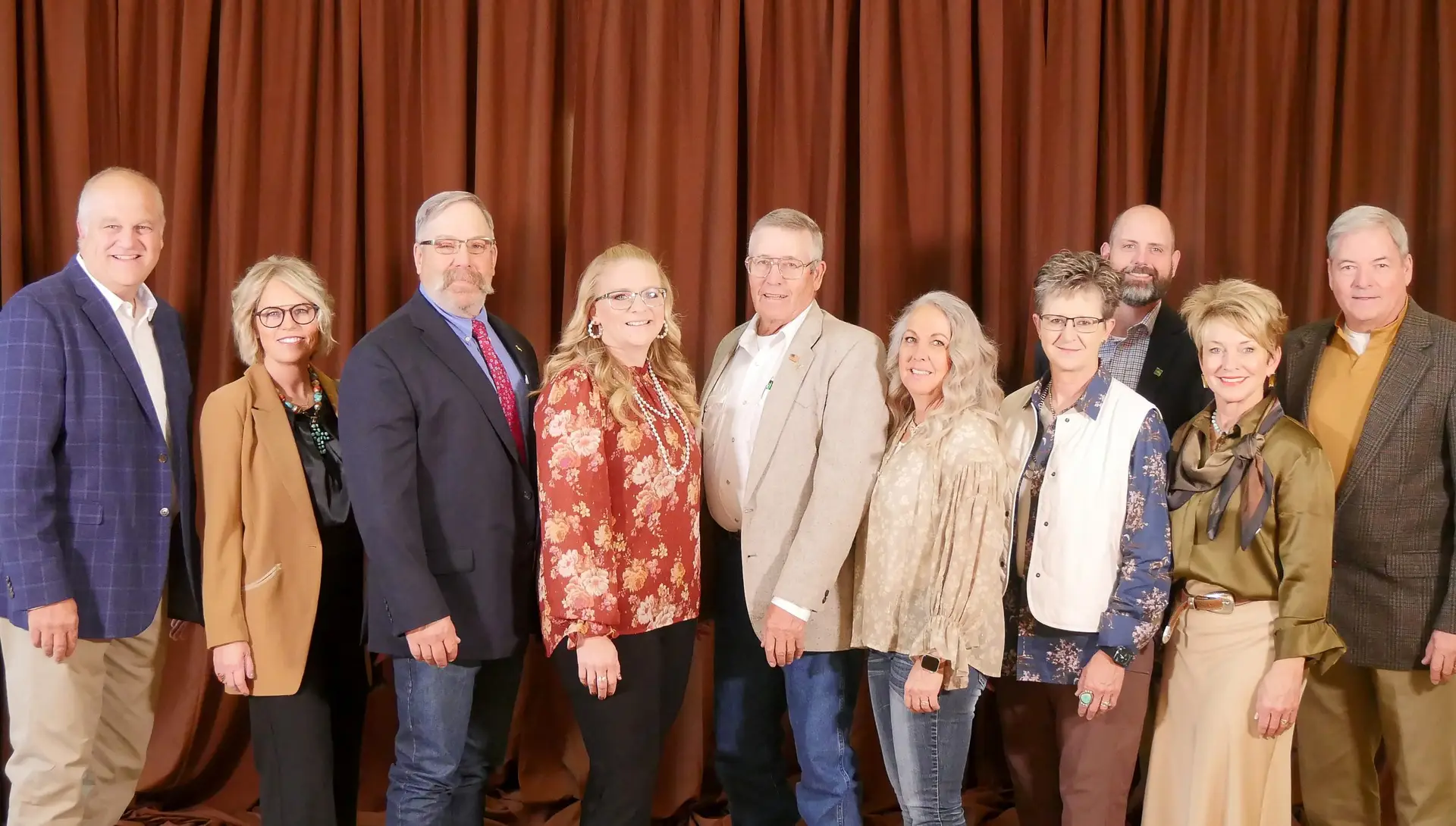 ten people wearing formal attire stand together in a line in front of a brown backdrop