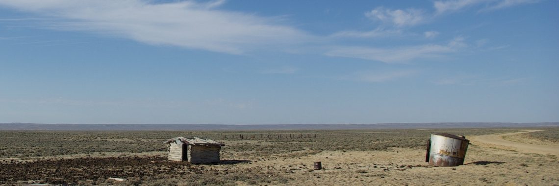 Parched landscape with scrubby shrubs, a tiny tumbledown cabin-like structure, and rusted metal cylinder under a blue sky. A dirt road curves through the landscape at right