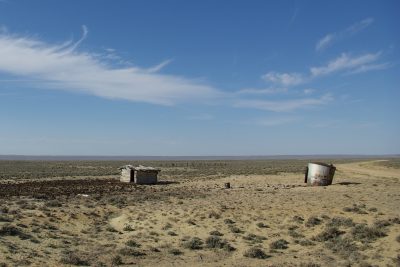 Parched landscape with scrubby shrubs, a tiny tumbledown cabin-like structure, and rusted metal cylinder under a blue sky. A dirt road curves through the landscape at right