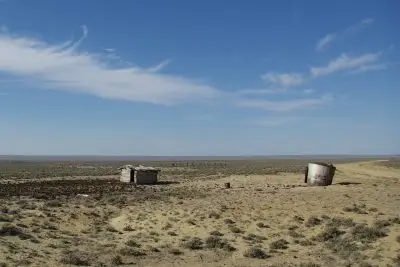 Parched landscape with scrubby shrubs, a tiny tumbledown cabin-like structure, and rusted metal cylinder under a blue sky. A dirt road curves through the landscape at right