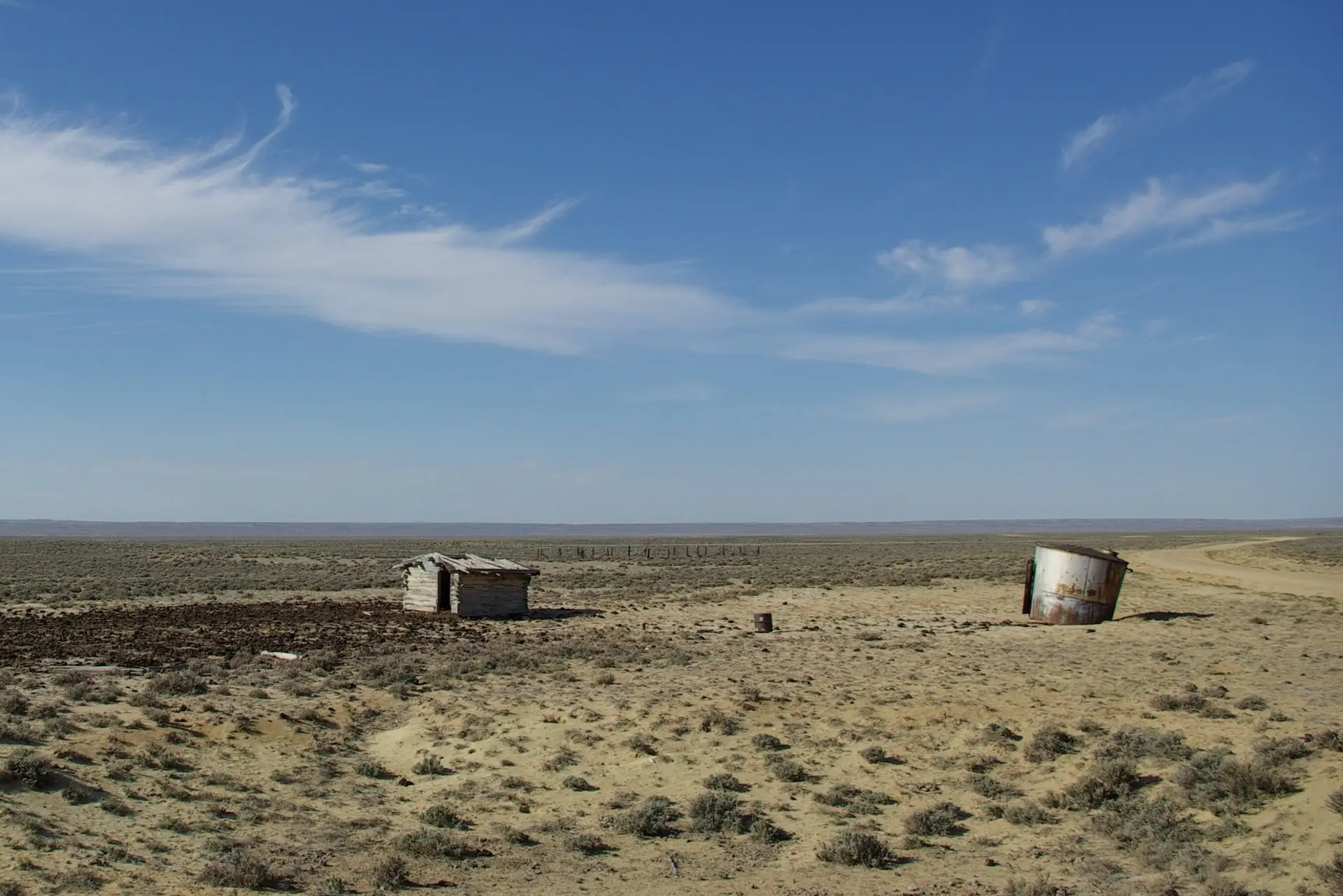 Parched landscape with scrubby shrubs, a tiny tumbledown cabin-like structure, and rusted metal cylinder under a blue sky. A dirt road curves through the landscape at right