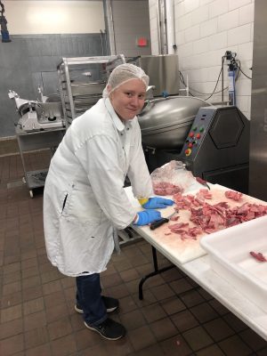 A young woman wearing a lab coat and gloves cuts pieces of meat.