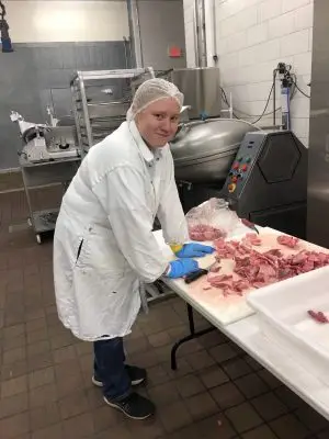 A young woman wearing a lab coat and gloves cuts pieces of meat. 