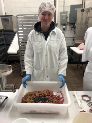A young woman wearing a lab coat and gloves holds a tray that contains ground meat with blueberries on top.