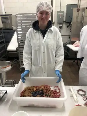 A young woman wearing a lab coat and gloves holds a tray that contains ground meat with blueberries on top. 