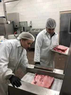 Two young women in lab coats hold trays with slices of meat.