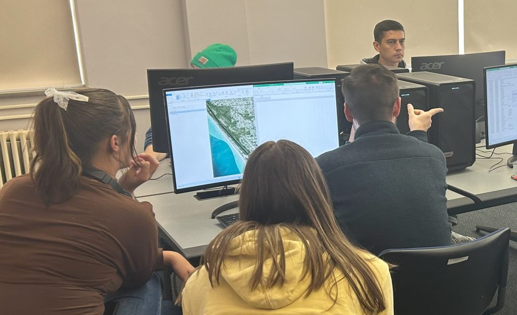 two woman and a man sit beside each other facing a monitor showing an aerial image of the Florida coastline. The man points his right hand toward another monitor.