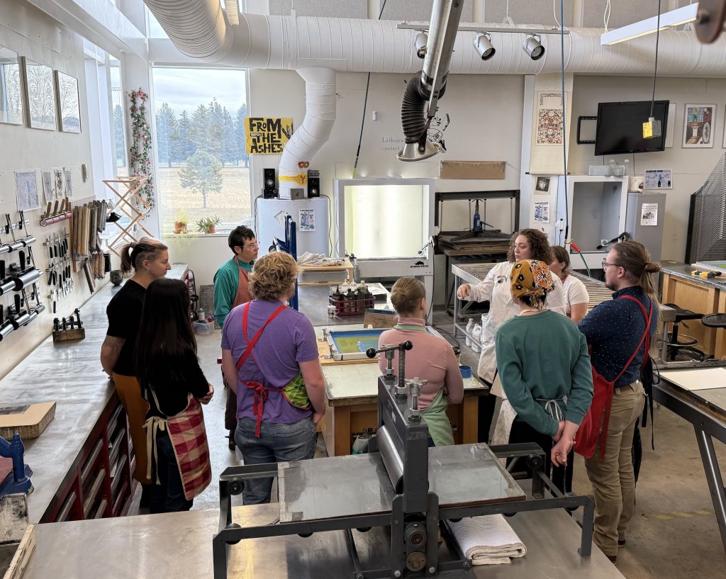 group of college students and their instructor stand in a spacious art studio, gathered around a table with a screen-printing screen on it