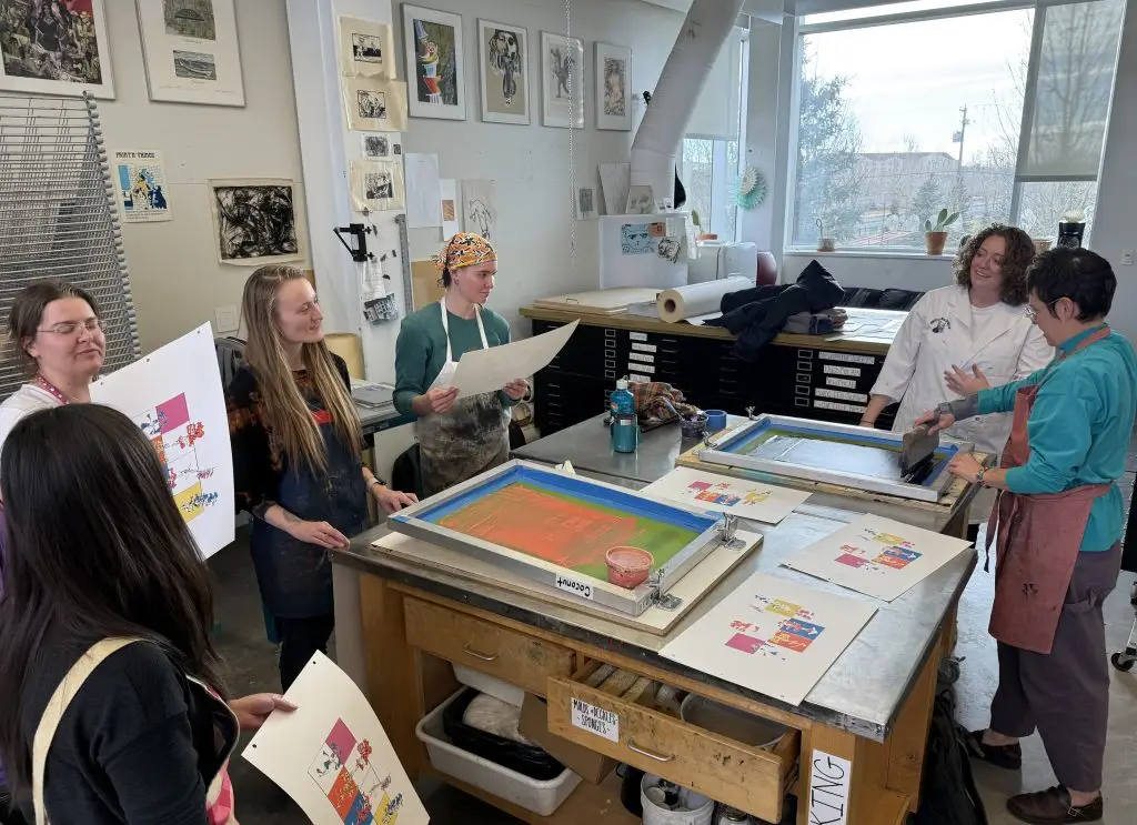 five college students and an instructor in an art studio stand around a table with screen-printing screens and prints on it. Several students hold colorful completed prints.