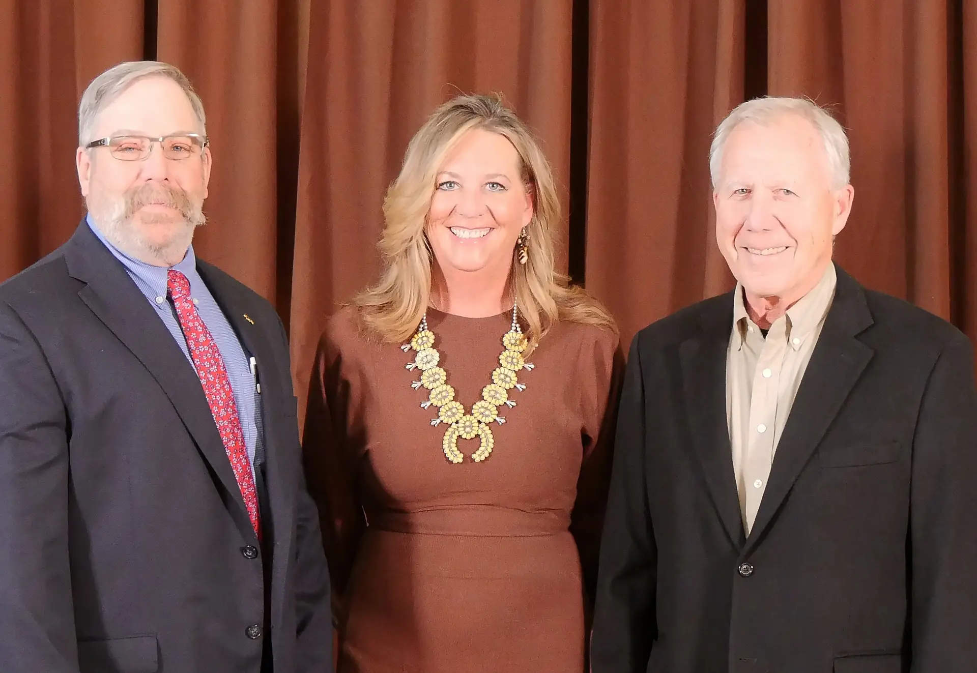 three smiling people wearing formal attire stand together in front of a brown backdrop