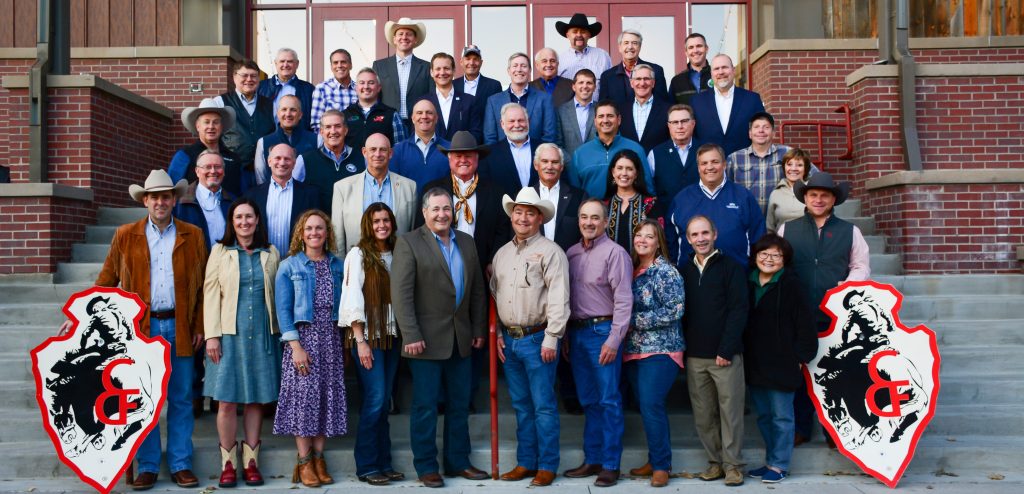 Large group of people wearing formal attire stand in rows on concrete steps in front of a building. On either side are arrowhead-shaped signs with black and red graphics of a cowboy on a bucking horse on a white background