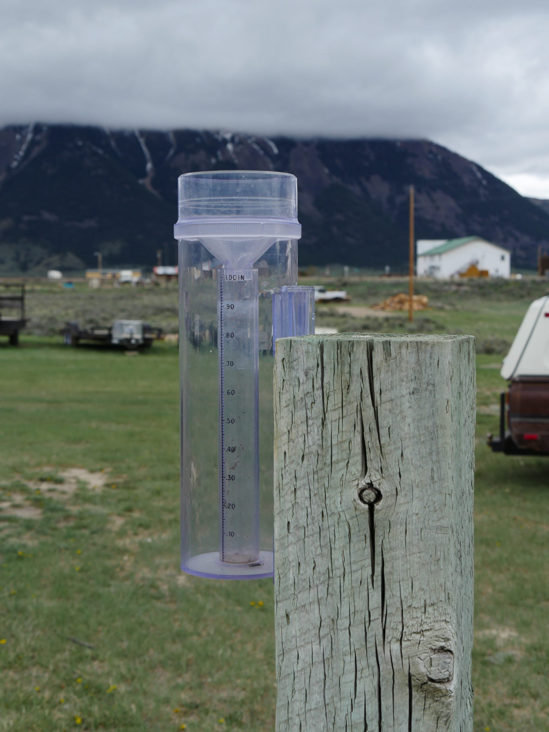 close-up of a rain gauge affixed to a wooden fence post with ranching equipment and a distance house blurred in the background. Beyond the house is a dark mountain shrouded in a low whitish gray cloud