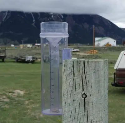 close-up of a rain gauge affixed to a wooden fence post with ranching equipment and a distance house blurred in the background. Beyond the house is a dark mountain shrouded in a low whitish gray cloud