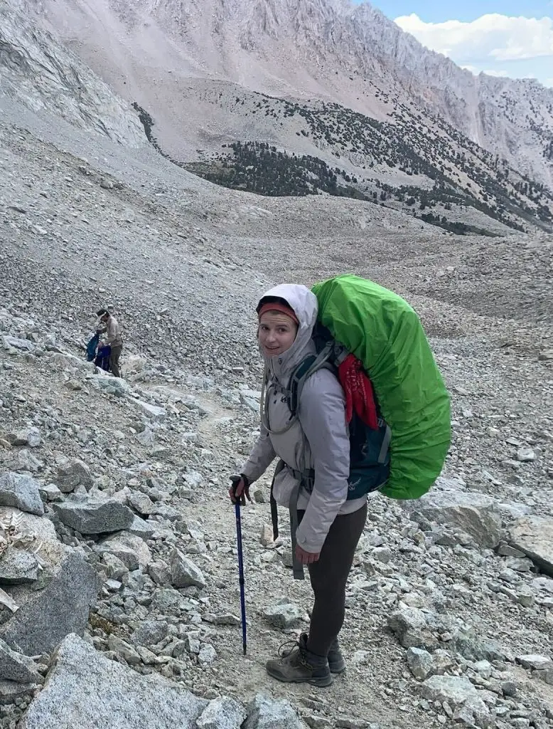 Woman wearing outdoor gear and carrying a backpacking backpack uses a trekking pole on a steep descent down a hillside with lots of loose rock. Farther down the slope, another figure adjusts the contents of his backpack