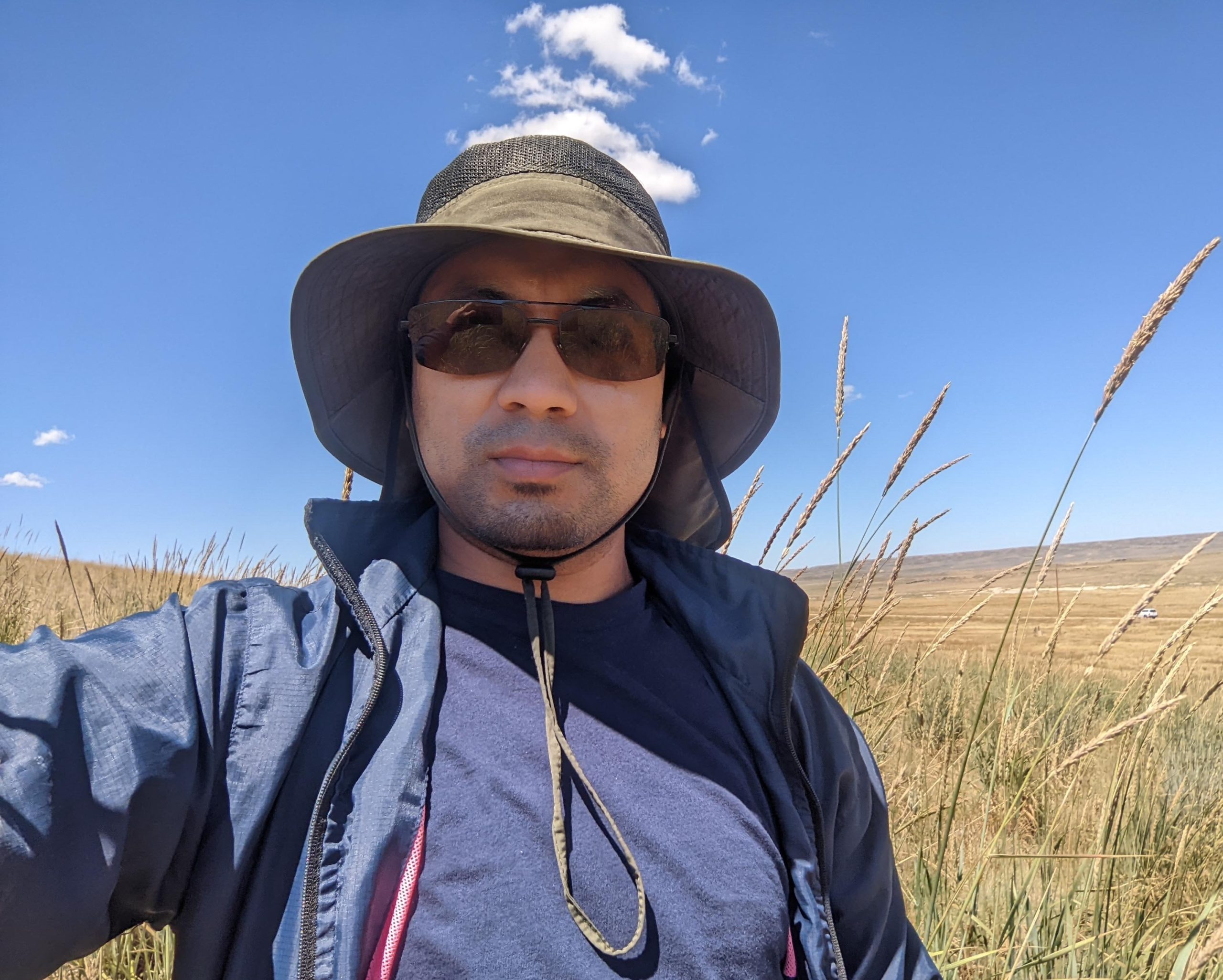 Selfie of a man wearing sun hat, sun glasses, and a windbreaker while standing in a field of tall yellow grass