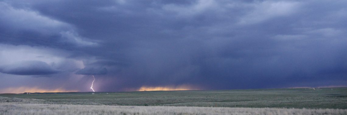 Looking out across a sagebrush plain, jagged white lightning bolt strikes the ground on the horizon underneath dark blue clouds tinged with peach