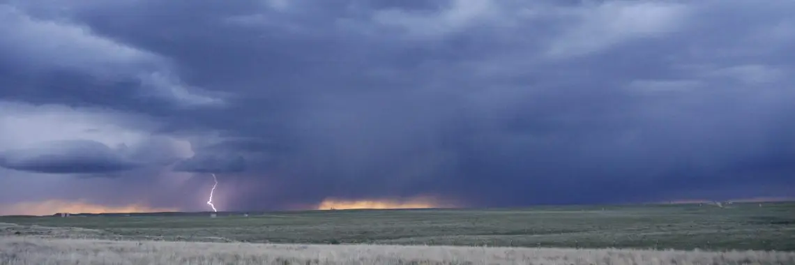 Looking out across a sagebrush plain, jagged white lightning bolt strikes the ground on the horizon underneath dark blue clouds tinged with peach