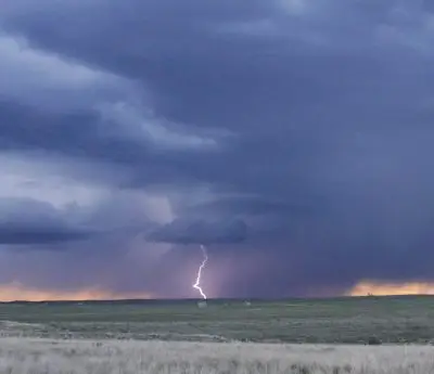 Looking out across a sagebrush plain, jagged white lightning bolt strikes the ground on the horizon underneath dark blue clouds tinged with peach