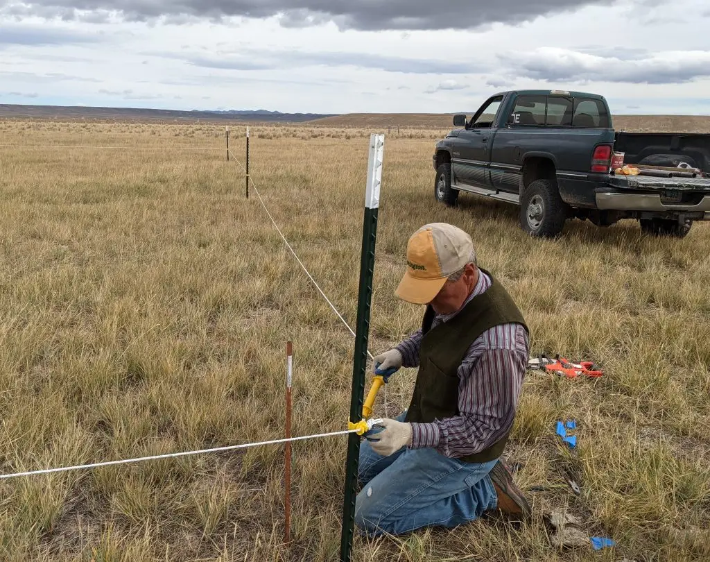 Man kneels in a grassy field to adjust a rope attached to metal poles to fence off a field site. A pickup truck with tools is parked nearby and dark clouds dot the sky overhead