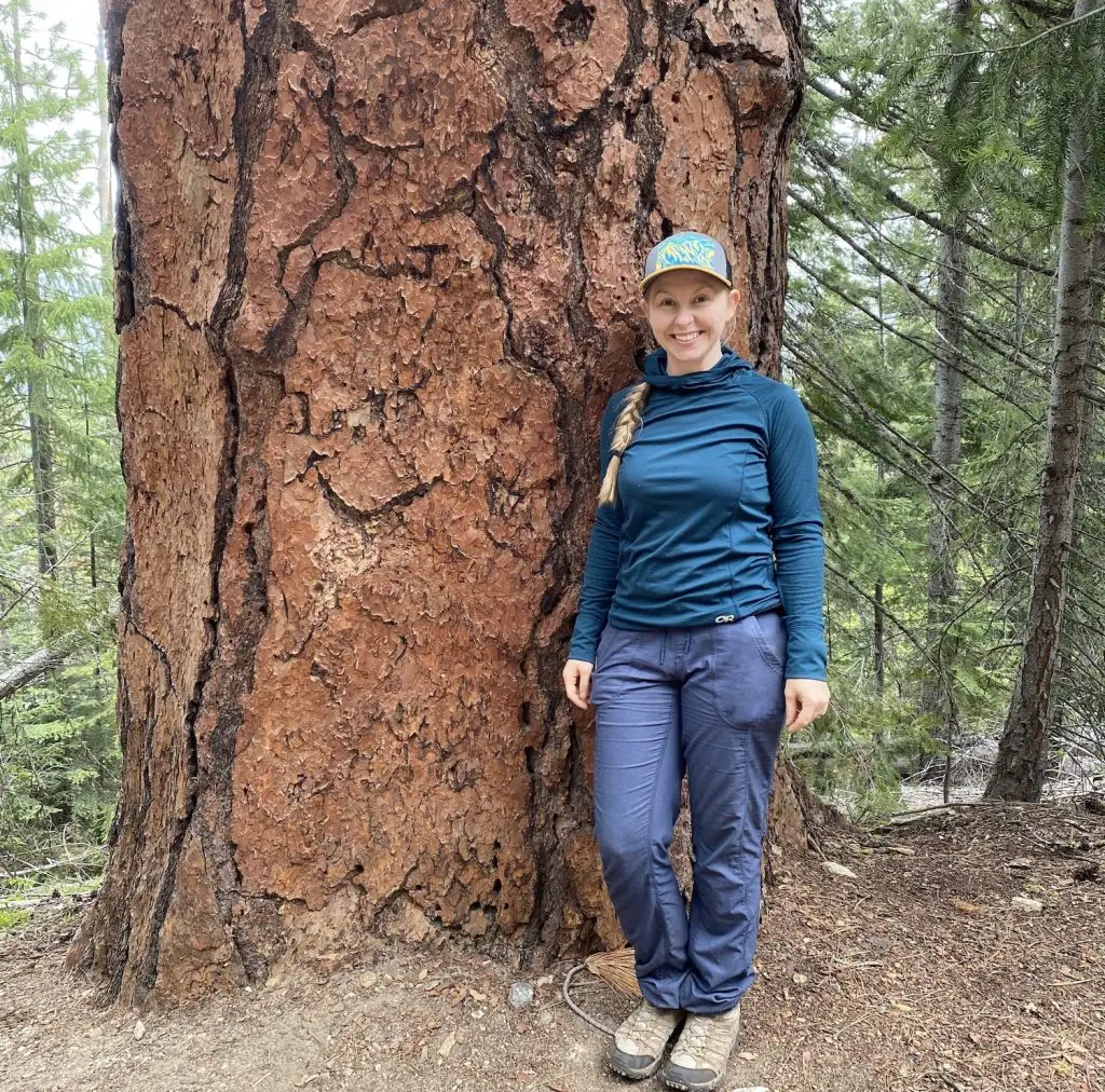 smiling woman wearing baseball cap and outdoor clothing stands in front of a giant pine tree in a forest