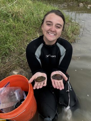 A woman holding out two mussels while kneeling by the shore of a shallow body of water.