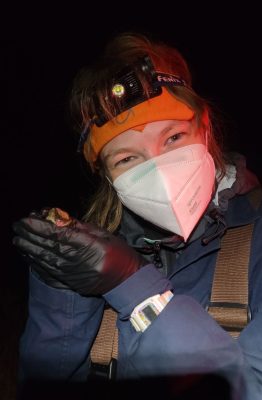 A woman wearing a headlamp, gloves, a mask, and warm clothing holding a bat.