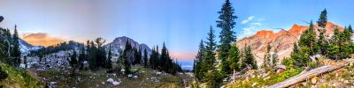 panorama of mountainous landscape dotted with dark fir trees, rocks and snowy crags with blue and pink sky above