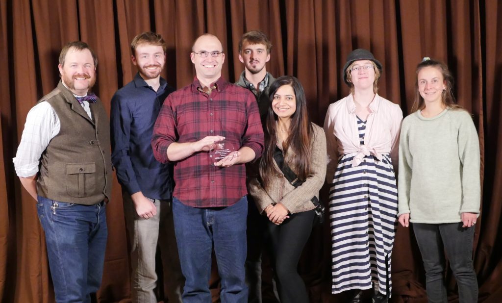 A group of people in semi-formal clothing. The person in the center has a small glass plaque.
