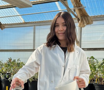 smiling woman wearing lab coat waters plants in a greenhouse using a glass beaker