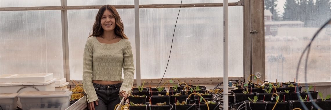 Drea Hineman stands in a greenhouse next to trays of plants connected to an irrigation system