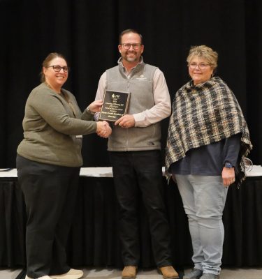 Three people in semi-formal clothing. The man in the middle is holding a plaque.