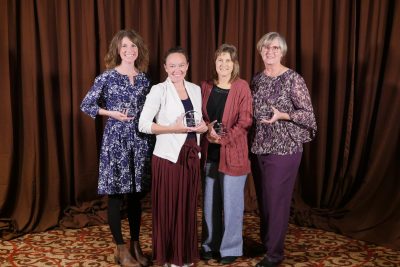 Four women in formal clothing each holding a small glass plaque.