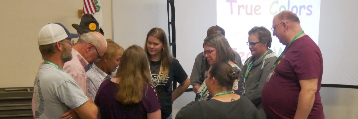 group of adults stand in a close circle with an American flag, 4-H flag, and screen labeled "True Colors" in the background.