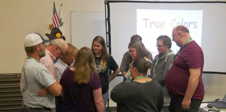 group of adults stand in a close circle with an American flag, 4-H flag, and screen labeled "True Colors" in the background.