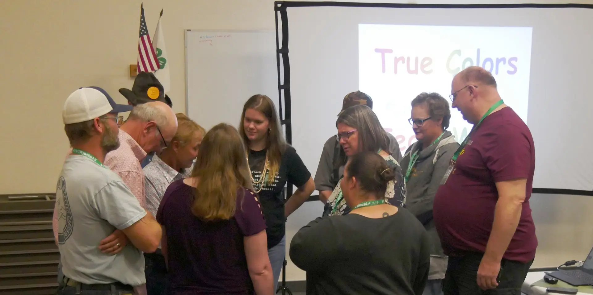 group of adults stand in a close circle with an American flag, 4-H flag, and screen labeled "True Colors" in the background.