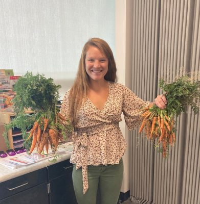CNP educator smiles at the camera, holding a large bunch of unwashed carrots by their leafy green stems in each hand
