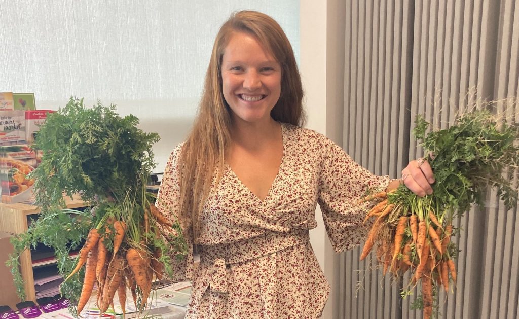 CNP educator smiles at the camera, holding a large bunch of unwashed carrots by their leafy green stems in each hand