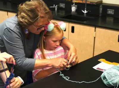 Woman teaches young girl how to hold knitting needles, guiding her hands as the yarn unwinds from a ball on the table in front of them