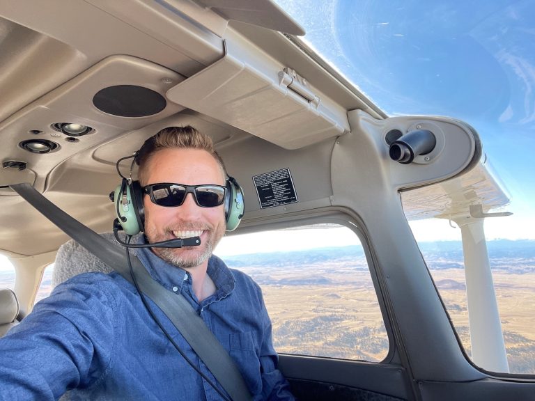 Christopher Robertson sits inside a small aircraft cockpit wearing a headset and sunglasses, smiling while flying over a rural landscape below.