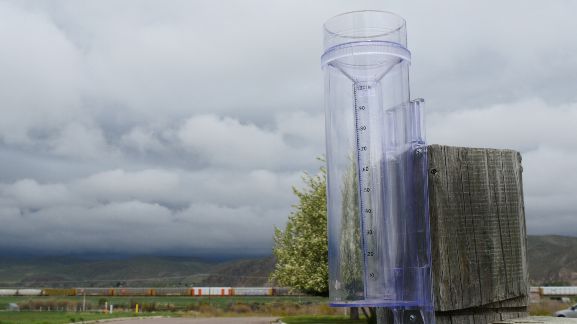 plastic precipitation gauge with measurements in inches affixed to a wooden post with mountains and a train in the background