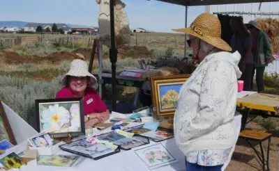 Artist booth at an Ag & Art Tour event, where a local artist displays watercolor paintings and handmade prints while engaging with a visitor; set outdoors with rural landscape, showcasing the connection between agriculture, creativity, and community.