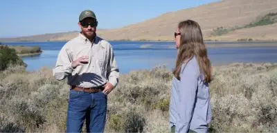 Two land managers stand in sagebrush near a lake and talk, with one gesturing while explaining something; rolling hills and water are visible in the background.
